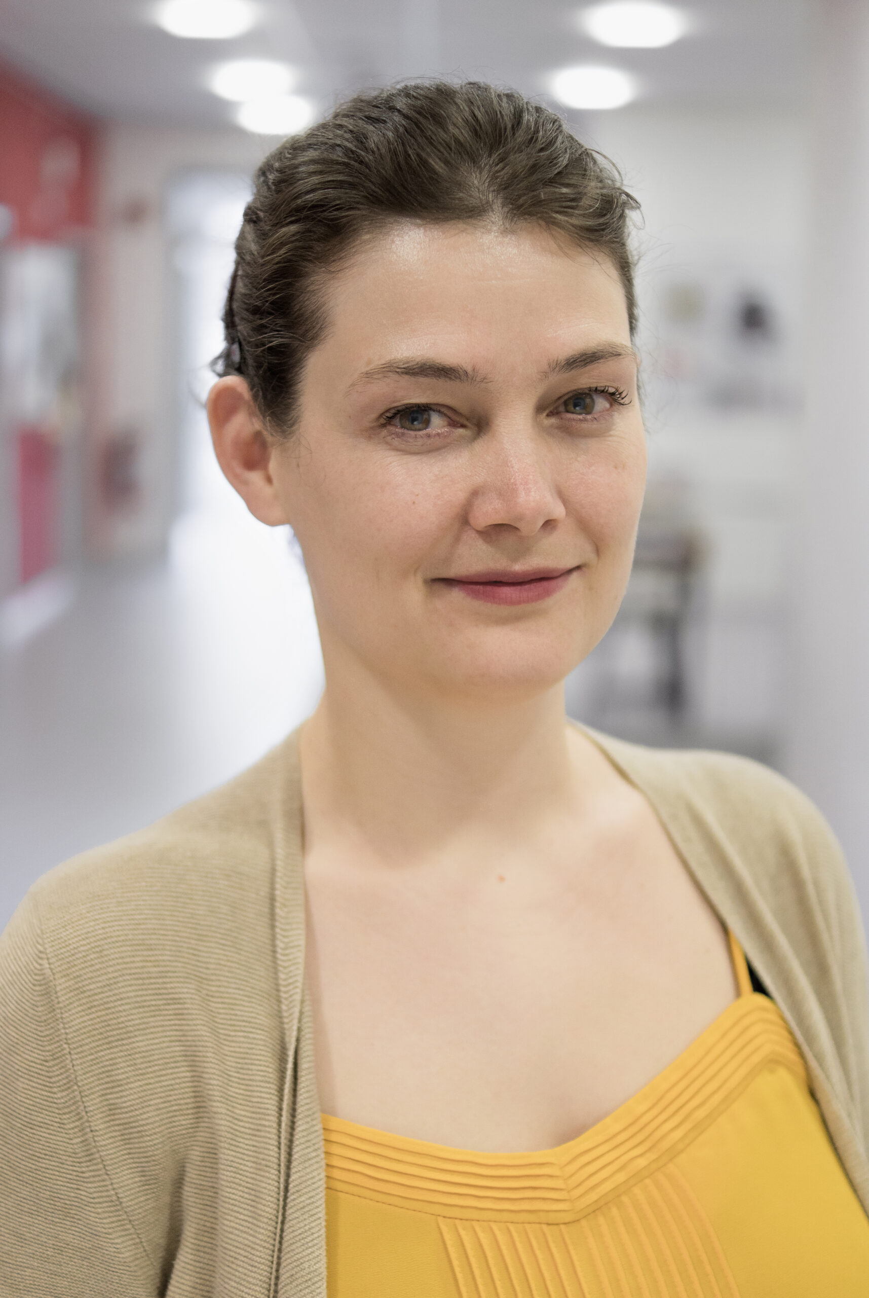 Eine Person mit kurzen braunen Haaren, einer beigen Strickjacke und einem gelben Oberteil steht in einem gut beleuchteten Flur der Hochschule Coburg. Sie lächelt sanft in die Kamera, der Hintergrund ist leicht unscharf.