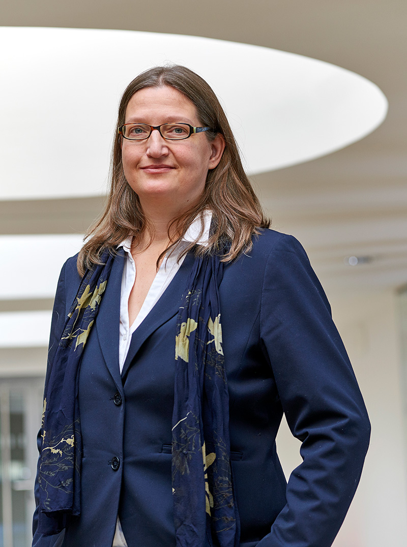 Eine Person mit langen Haaren, Brille, blauem Anzug, weißem Hemd und marineblauem Schal steht selbstbewusst im modernen Innenraum der Hochschule Coburg, beleuchtet von einem kreisförmigen Lichtelement darüber.