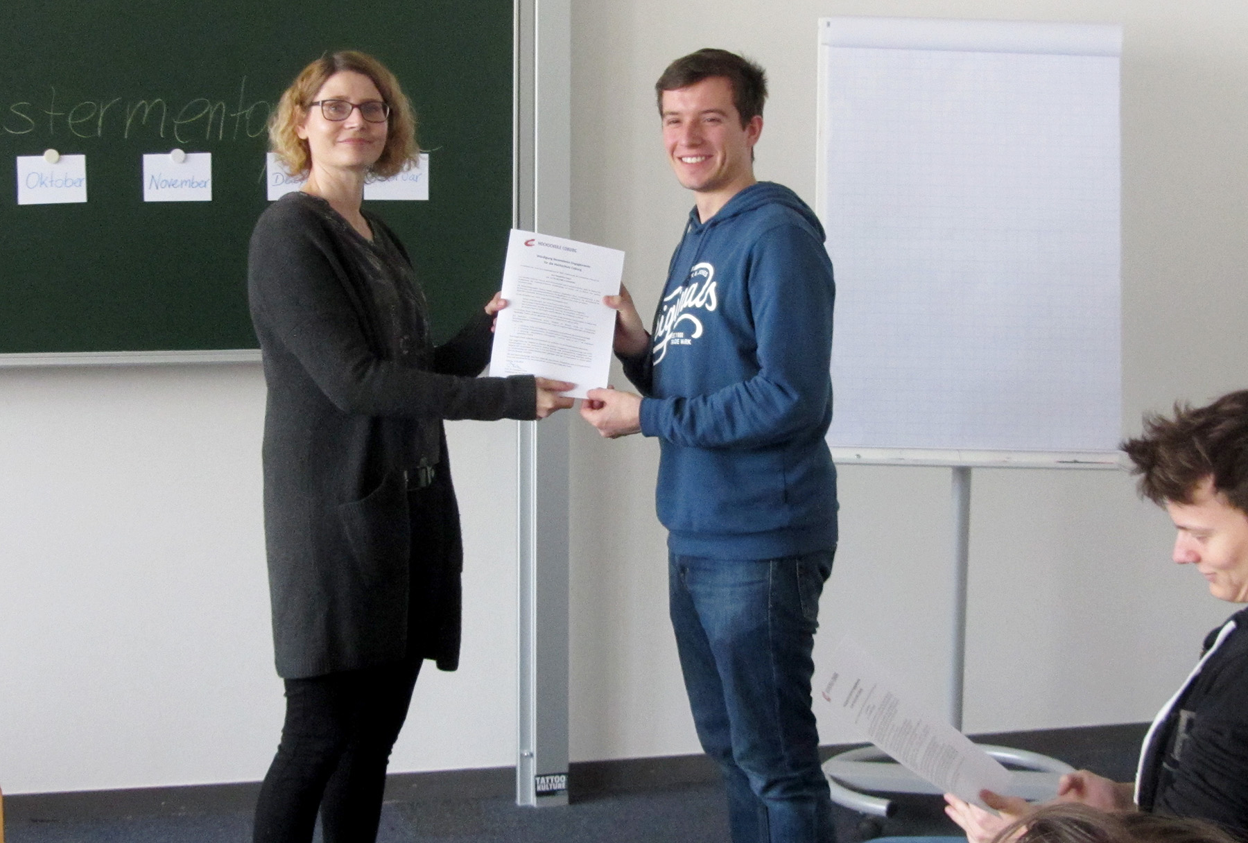 Eine Frau der Hochschule Coburg überreicht einem lächelnden Mann in einem blauen Kapuzenpullover in einem Klassenzimmer ein Zertifikat. Im Hintergrund sind eine grüne Tafel mit Papieren und ein Flipchart zu sehen. Eine weitere Person sitzt in der Nähe und betrachtet ein Dokument.