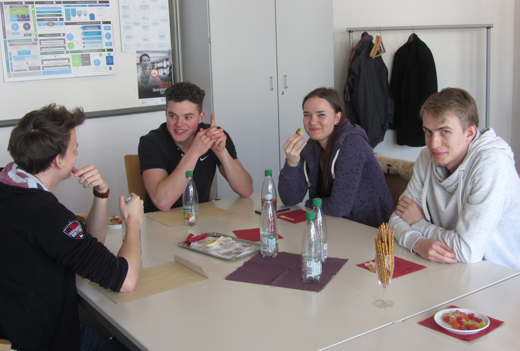 Vier junge Erwachsene sitzen an einem Tisch in der Hochschule Coburg und genießen einen gemütlichen Snack. Auf dem Tisch stehen Wasserflaschen, Brezeln und Teller mit Essen. Sie lächeln und scheinen in ein Gespräch vertieft zu sein. Im Hintergrund sind eine Garderobe und ein helles Poster zu sehen.