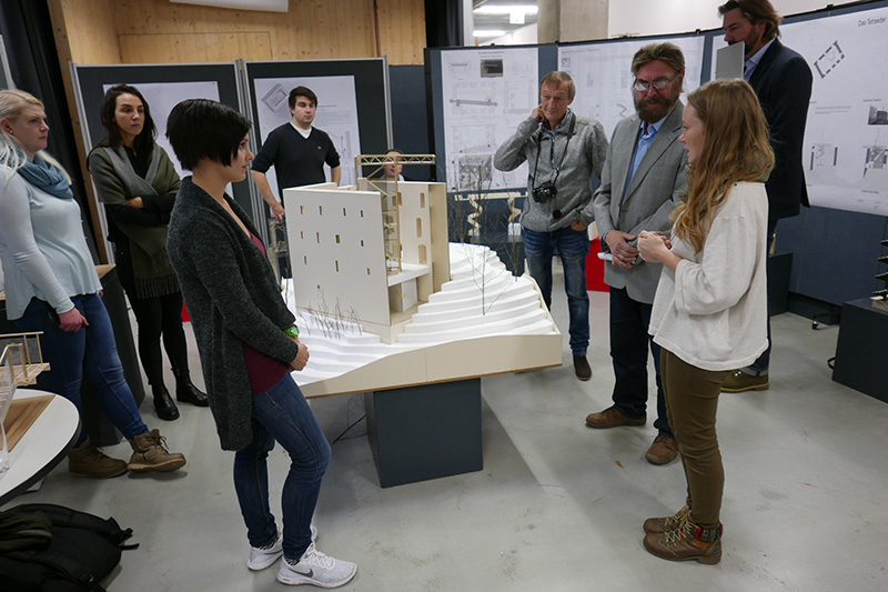 Eine Gruppe von Menschen versammelt sich um ein großes Architekturmodell auf einem Tisch in der Hochschule Coburg. Im Hintergrund des Raums sind auf Tafeln Projektpräsentationen zu sehen. Einige machen sich Notizen, andere betrachten und diskutieren das Modell mit großem Interesse.