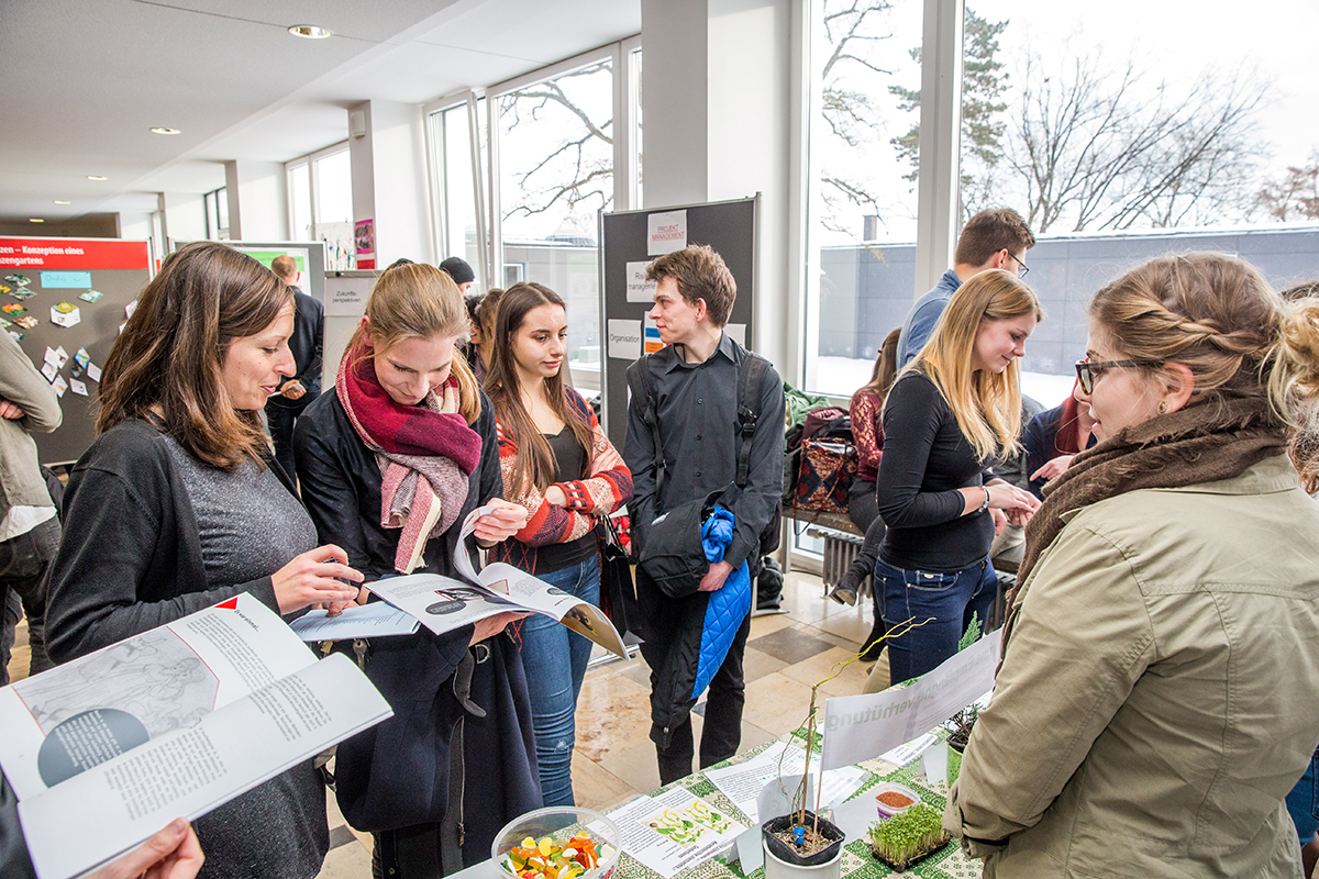 In einer hellen, modernen Wissenschaftsausstellung an der Hochschule Coburg beschäftigen sich Menschen mit kleinen Pflanzen und Informationsmaterialien. Einige lesen Broschüren, während andere angeregt über die Exponate diskutieren. Und das alles vor dem Hintergrund großer Fenster, die den Raum mit Licht durchfluten.