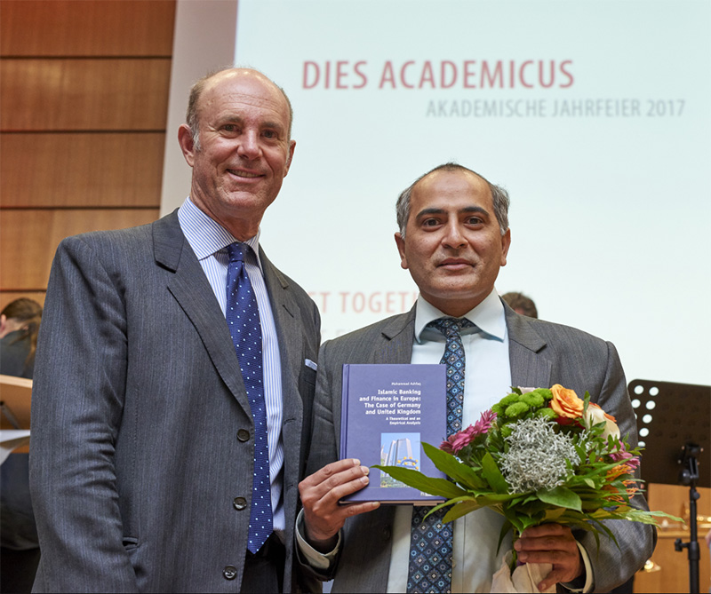 Zwei Männer in Anzügen stehen zusammen und lächeln herzlich. Einer hält ein Buch und einen Blumenstrauß in der Hand. Hinter ihnen ist auf einem Präsentationsbildschirm der Titel „Dies Academicus Akademische Jahresfeier 2017“ zu sehen, der den Geist der Hochschule Coburg feiert.