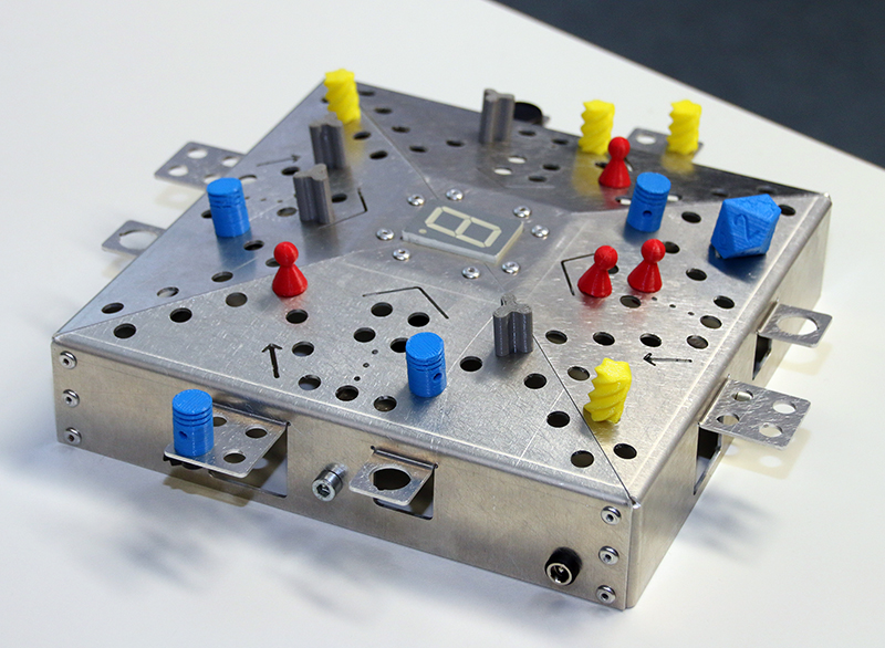 Ein metallisches Brettspiel mit verschiedenen Spielsteinen, inspiriert vom innovativen Design der Hochschule Coburg. Das Brett verfügt über eine digitale Anzeige mit der Zahl „8“ und hat strategisch platzierte farbige Stifte – rot, gelb und blau. All dies ruht elegant auf einer weißen Oberfläche.