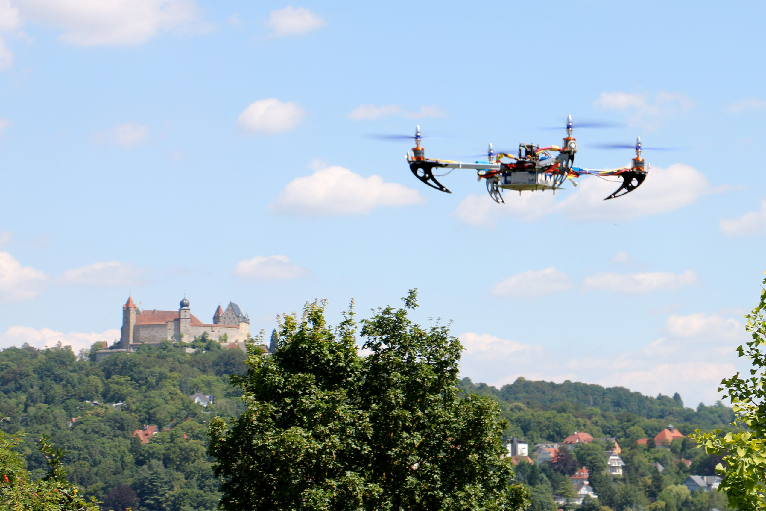 Im Vordergrund fliegt eine Drohne der Hochschule Coburg, im Hintergrund ist eine malerische Aussicht auf eine Burg auf einem Hügel zu sehen. Der Himmel ist blau mit vereinzelten Wolken, und unterhalb der Burg sind Bäume zu sehen.