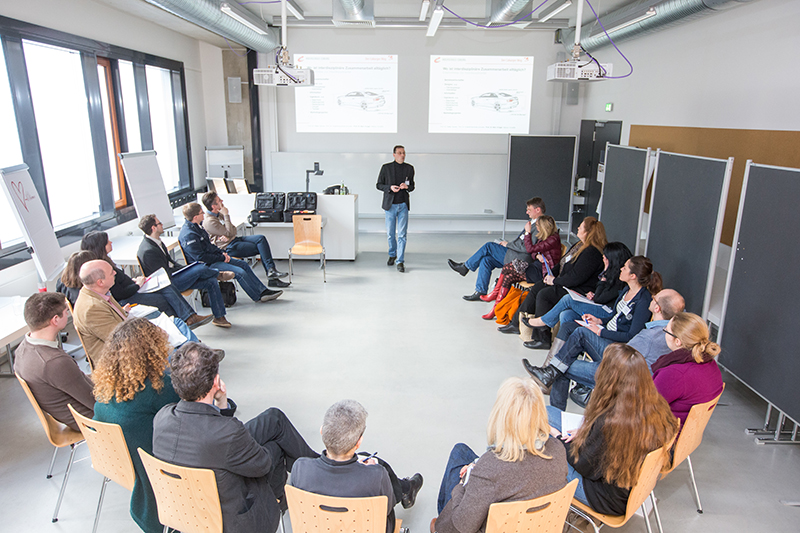 In einem hellen Unterrichtsraum der Hochschule Coburg sitzt eine Gruppe von Menschen im Kreis und blickt aufmerksam auf den Vortragenden. Hinter ihnen laufen auf zwei Projektionsflächen Folien, die der spannenden Präsentation Tiefe verleihen. Der Raum verfügt über helle Wände und Holzstühle, was eine Atmosphäre des Lernens und der Zusammenarbeit fördert.