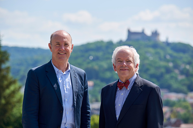 Zwei Männer in Anzügen stehen im Freien. Im Hintergrund sind ein Hügel und das historische Schloss der Hochschule Coburg zu sehen. Sie lächeln beide und der Himmel ist klar.