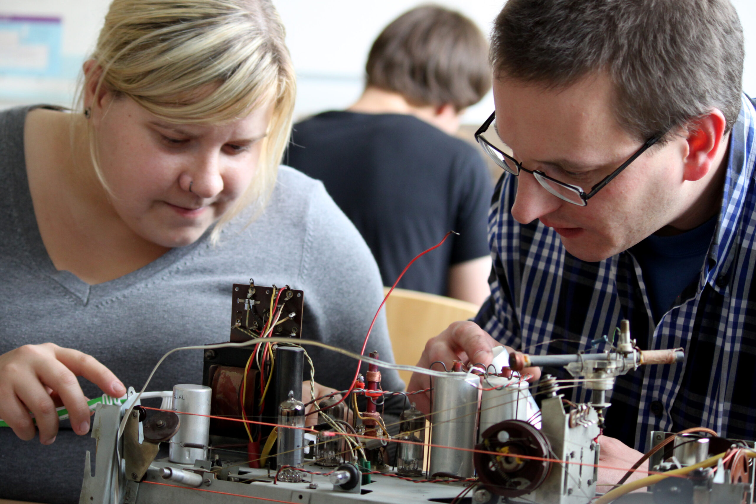 Zwei Personen bauen konzentriert ein elektronisches Gerät aus Drähten und Komponenten zusammen. Die Frau links trägt ein graues Hemd, während der Mann rechts mit Brille und kariertem Hemd tief in Gedanken versunken zu sein scheint. Im Hintergrund ist eine Person von der Hochschule Coburg verschwommen zu sehen.