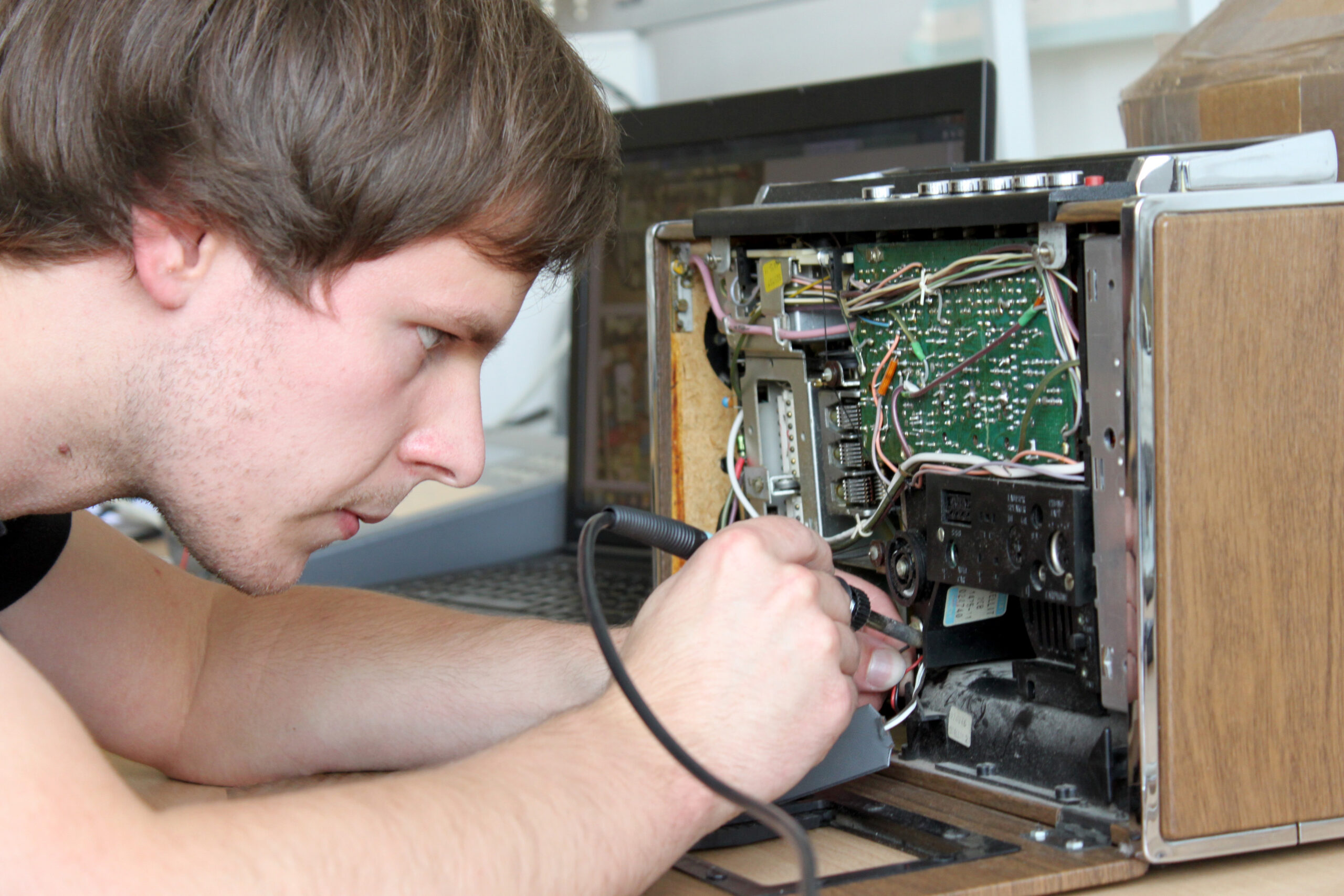 Eine Person lötet konzentriert mit einem Lötkolben die internen Schaltkreise eines alten elektronischen Geräts, möglicherweise repariert sie es für ein Projekt an der Hochschule Coburg. Die Holzaußenseite des Geräts ist teilweise geöffnet, sodass Kabel und Komponenten sichtbar sind. Im Hintergrund ist ein Laptop zu sehen.