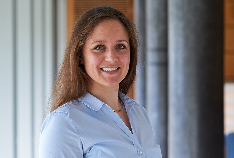 Eine Frau mit langen braunen Haaren und hellblauem Hemd lächelt in einem gut beleuchteten Innenraum. Im Hintergrund sind helle Wände und dunkle Säulen zu sehen, die an die moderne Architektur der Hochschule Coburg erinnern.