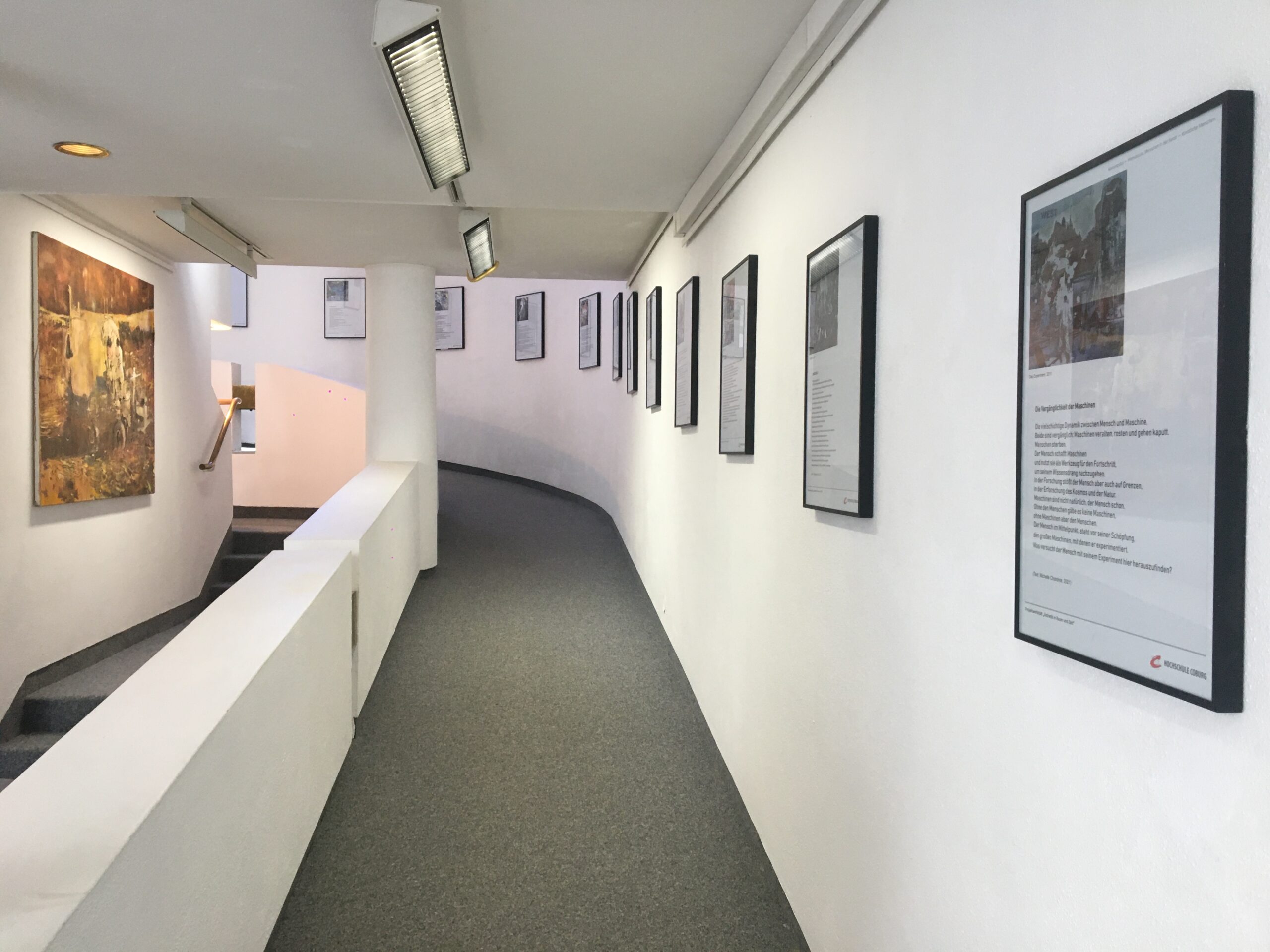 Eine moderne Galerie an der Hochschule Coburg verfügt über eine geschwungene weiße Wand, an der gerahmte Kunstwerke ausgestellt sind. Gleichmäßig angeordnete Rahmen lenken den Blick auf eine Treppe im Hintergrund. Deckenleuchten beleuchten jedes Stück, während der Teppichboden das minimalistische Design unterstreicht und eine elegante Atmosphäre schafft.