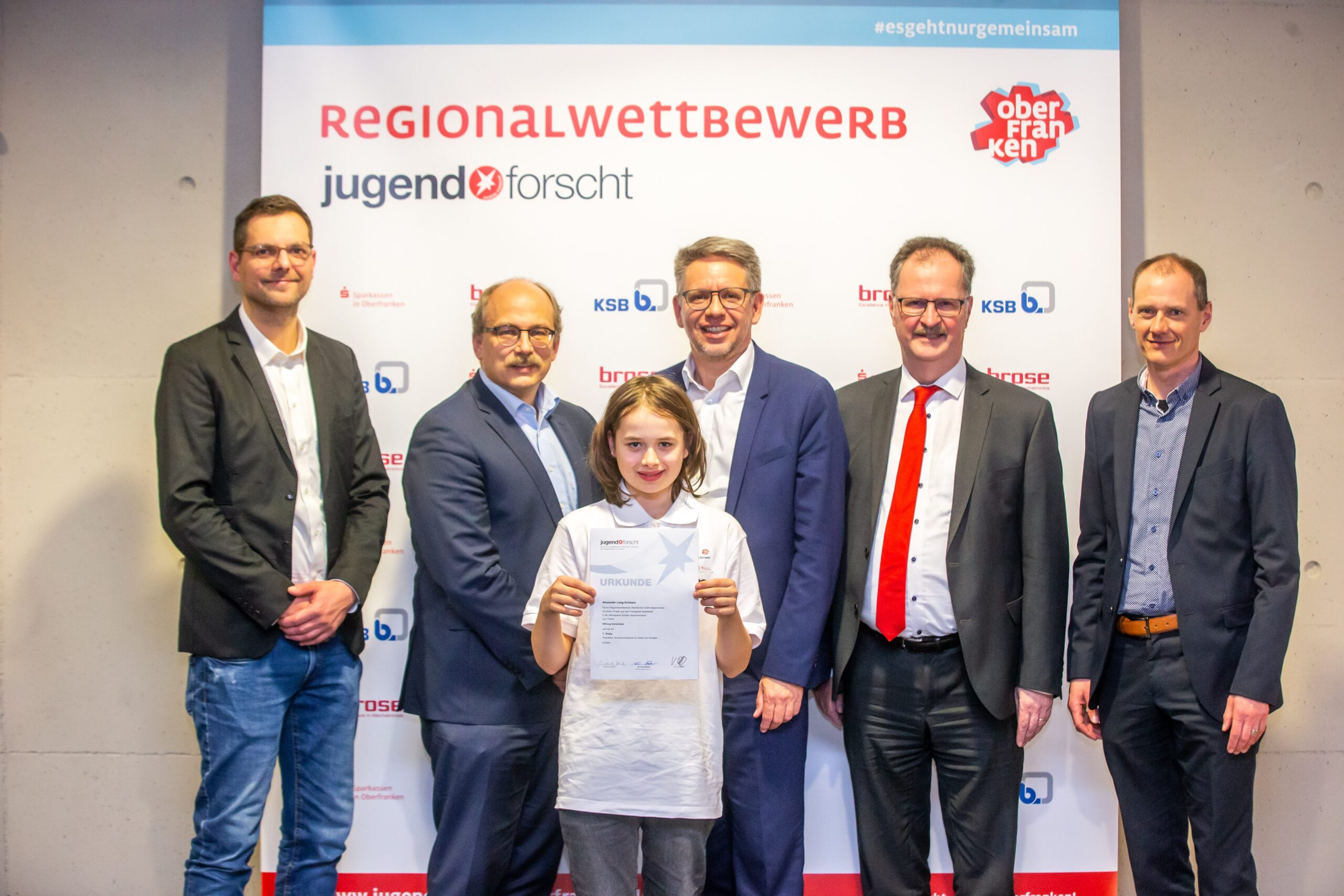 Ein junger Mensch steht stolz mit einer Urkunde vor einem Schild mit der Aufschrift „Regionalwettbewerb Jugend forscht Oberfranken“ an der Hochschule Coburg, umgeben von fünf Erwachsenen in Business-Kleidung, die für ein Gruppenfoto posieren.