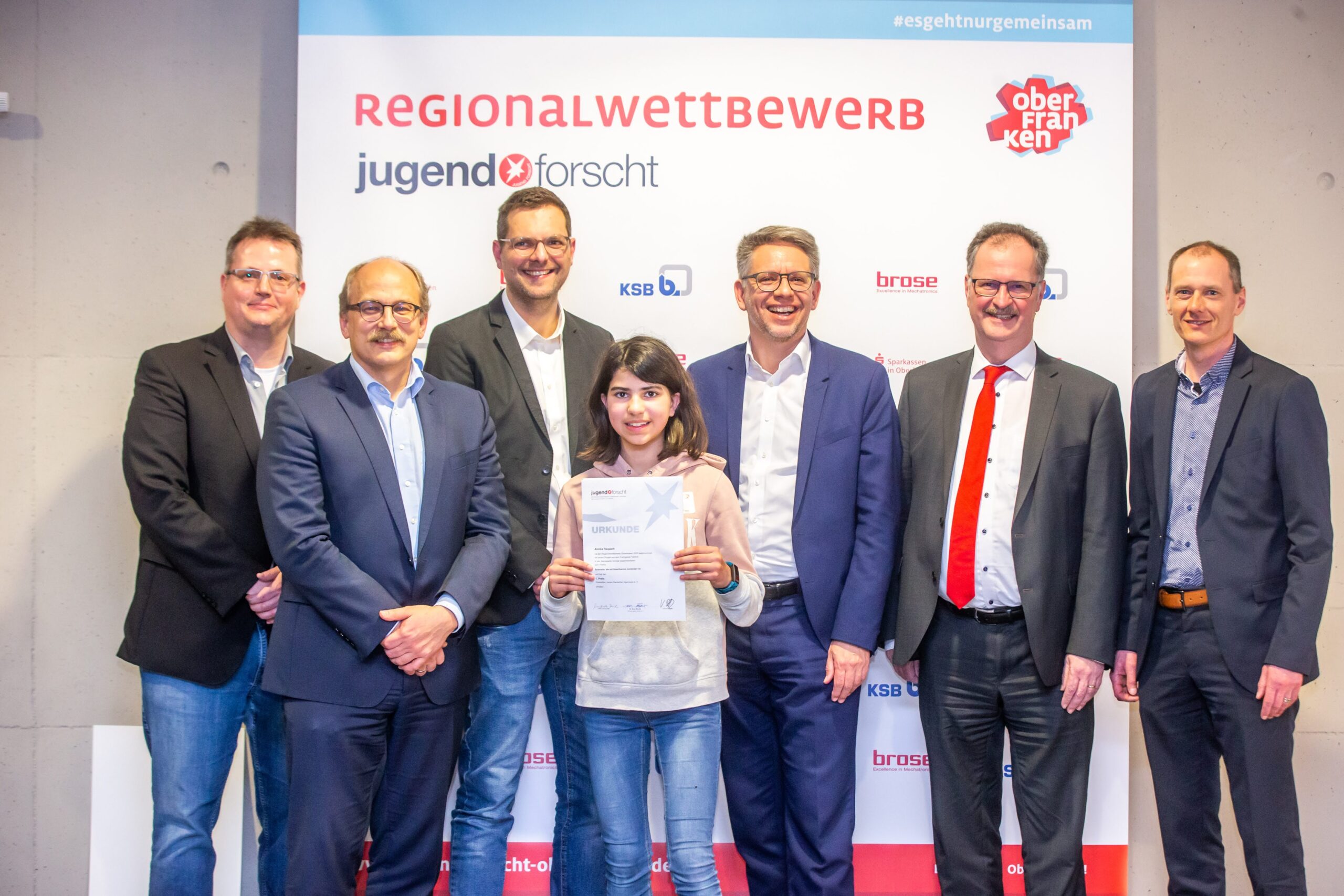 Ein junges Mädchen hält eine Urkunde in der Hand, während es mit mehreren Erwachsenen in Abendgarderobe an der Hochschule Coburg steht. Sie posieren für ein Gruppenfoto bei einer regionalen Wettbewerbsveranstaltung. Im Hintergrund ist ein Banner mit Logos und Text zu sehen, das den Geist des prestigeträchtigen Anlasses einfängt.