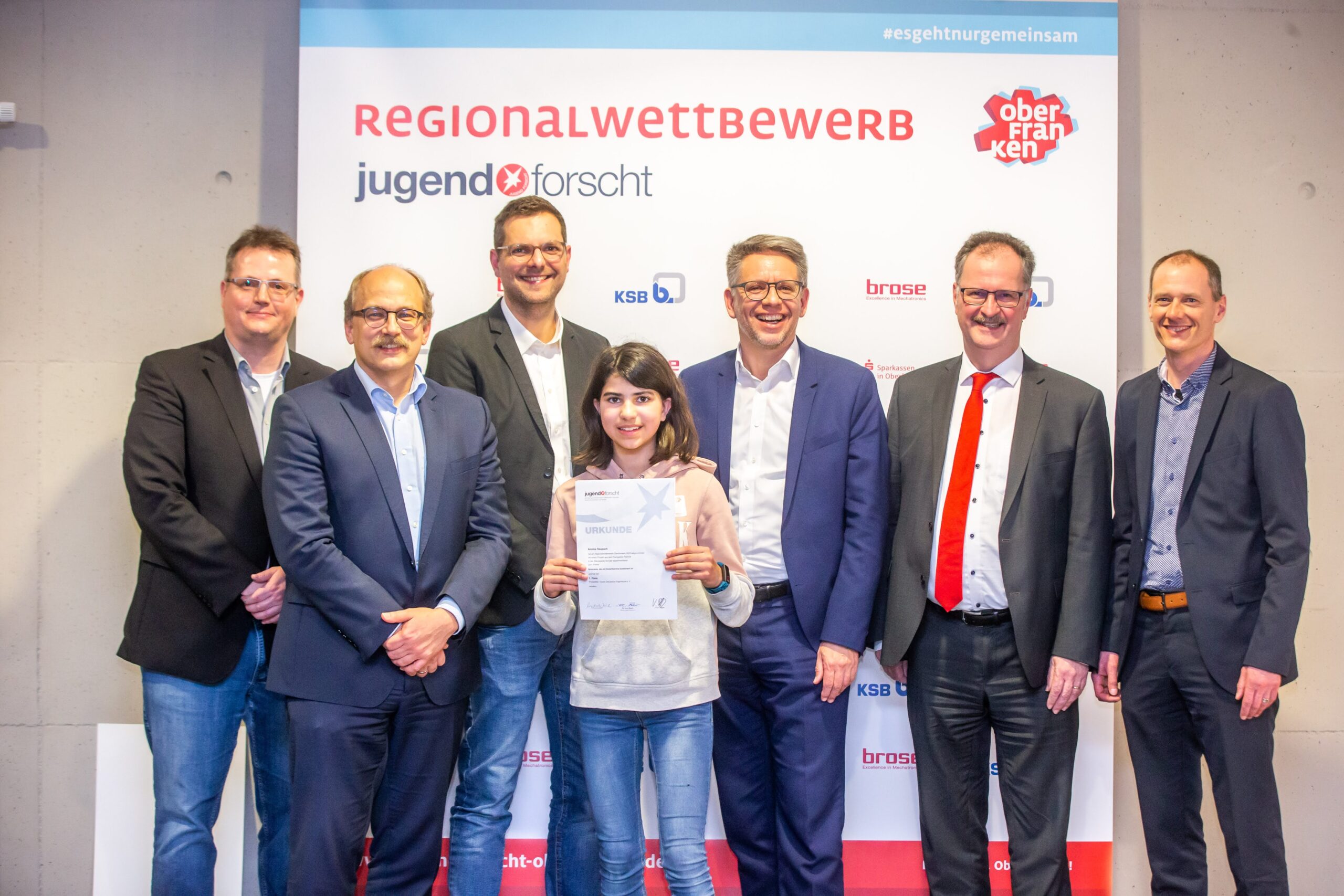 Eine Gruppe von acht Personen steht lächelnd vor einem Banner mit der Aufschrift „Regionalwettbewerb Jugend forscht“ an der Hochschule Coburg. Eine Person in der ersten Reihe hält eine Urkunde in der Hand. Im Hintergrund sind Logos und deutscher Text zu sehen.