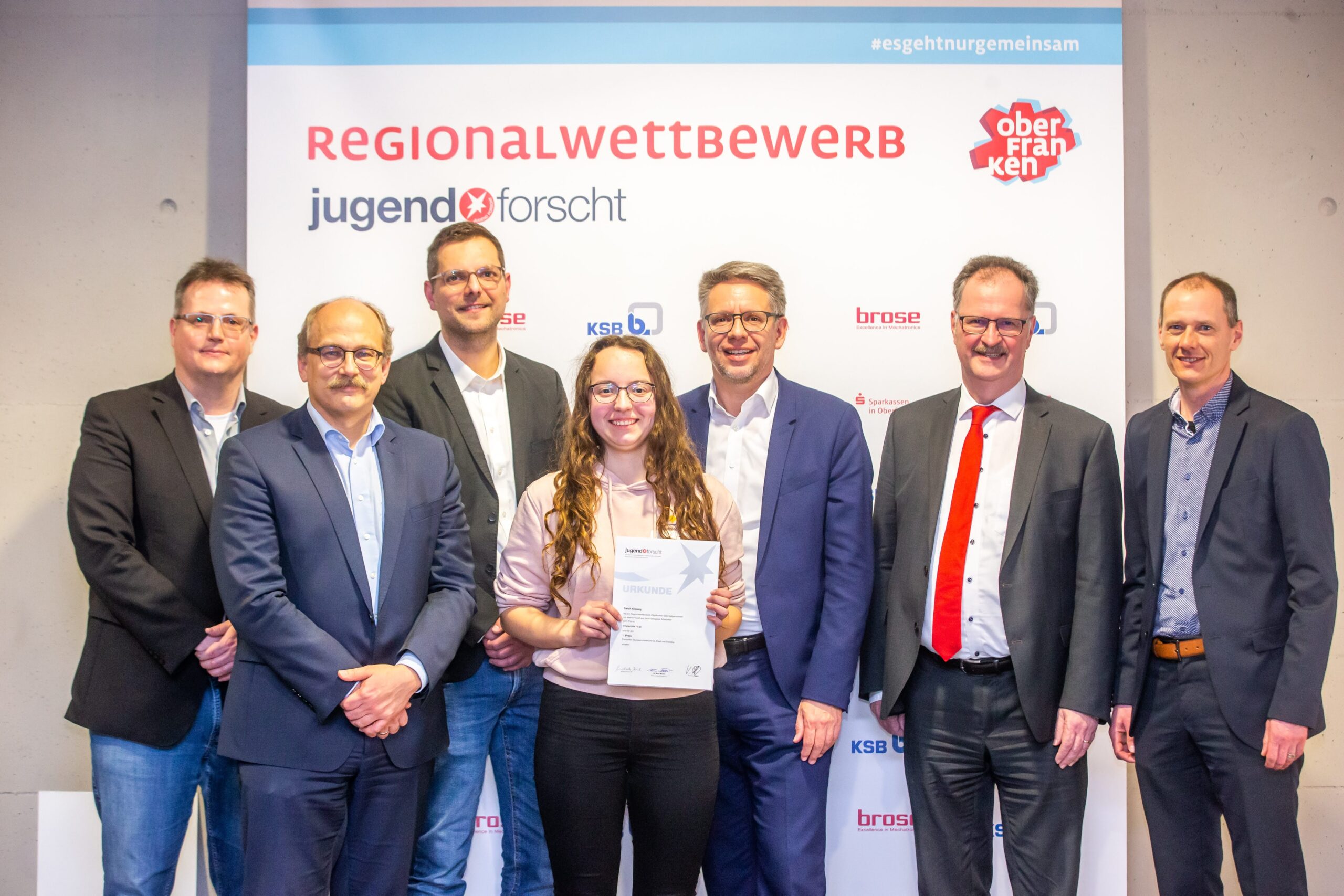 Eine Gruppe von sieben Personen in formeller Kleidung steht stolz vor einem Hintergrund mit der Aufschrift „Regionalwettbewerb Jugend forscht“. Sie lächeln alle, und die Person in der Mitte hält eine Urkunde in der Hand. Sie feiern ihren Erfolg an der Hochschule Coburg und strahlen Triumph und Kameradschaft aus.