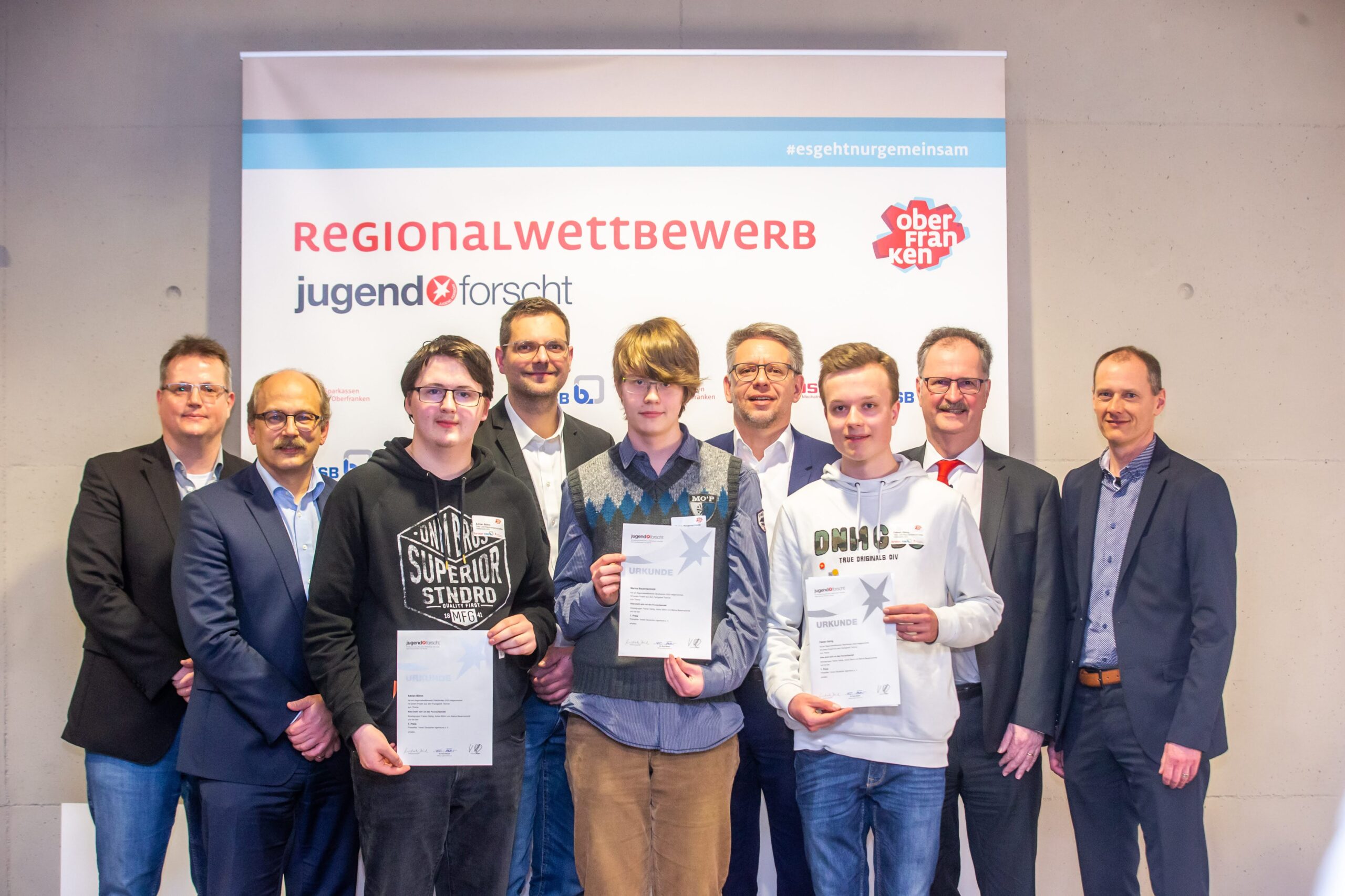 Eine Gruppe von Menschen steht in zwei Reihen. In der ersten Reihe stehen vier junge Menschen mit Zertifikaten, in der hinteren Reihe fünf Erwachsene in Anzügen. Über ihnen steht auf einem Banner „Regionalwettbewerb Jugend forscht Oberfranken“, gesponsert von der Hochschule Coburg.