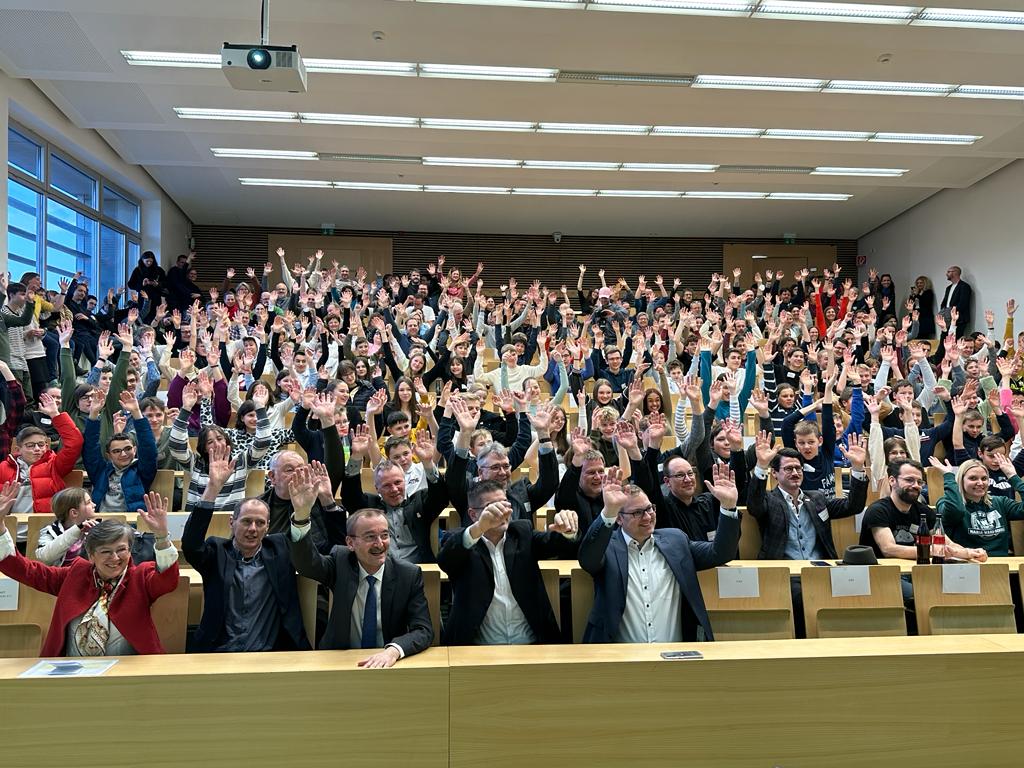 Eine große Gruppe von Menschen in einem Hörsaal der Hochschule Coburg posiert mit erhobenen Händen für ein Foto. Der Raum ist voller Reihen von Teilnehmern, und vorne stehen Tische, an denen mehrere Personen sitzen und in die Kamera lächeln.