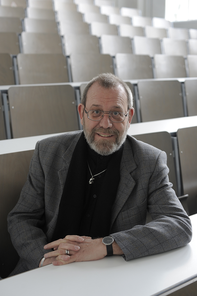 Ein bärtiger Mann mit Brille und grauem Anzug sitzt an einem weißen Schreibtisch in einem leeren Hörsaal mit Holzsitzen an der Hochschule Coburg. Mit gefalteten Händen blickt er in die Kamera.