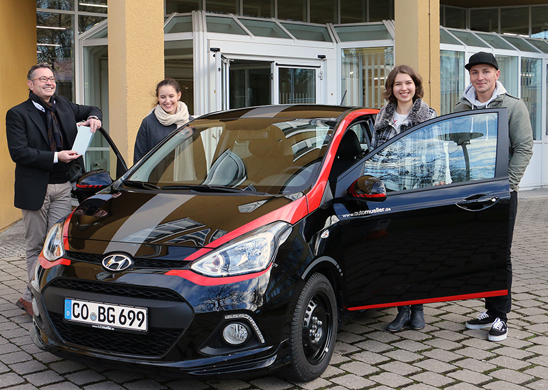 Vier Menschen stehen um einen schwarzen Hyundai mit roten Akzenten herum, zwei halten die Autotüren offen. Sie lächeln sich vor einem hellgelben Gebäude an, das an die lebendige Atmosphäre der Hochschule Coburg erinnert.