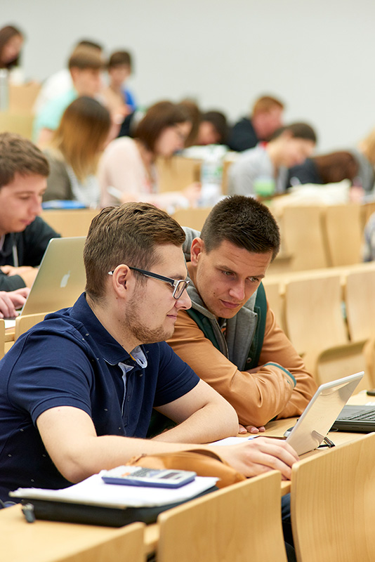 In einem Hörsaal der Hochschule Coburg sitzen Studierende mit Laptops und Notebooks an Tischen und diskutieren angeregt. Im Hintergrund sind weitere Studierende zu sehen, die in ihre Geräte und Notizen vertieft sind. So entsteht eine aufmerksame und zugleich akademische Atmosphäre.