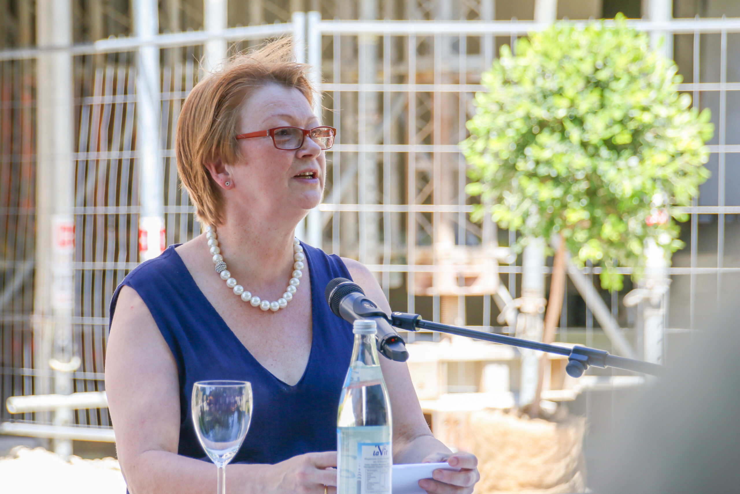 Eine Frau in einem blauen Kleid, Vertreterin der Hochschule Coburg, spricht bei einer Veranstaltung im Freien in ein Mikrofon. Sie trägt eine Brille und eine Perlenkette. Vor ihr stehen eine Wasserflasche und ein leeres Weinglas. Im Hintergrund verschwimmen sanft ein Baum und ein Metallzaun.