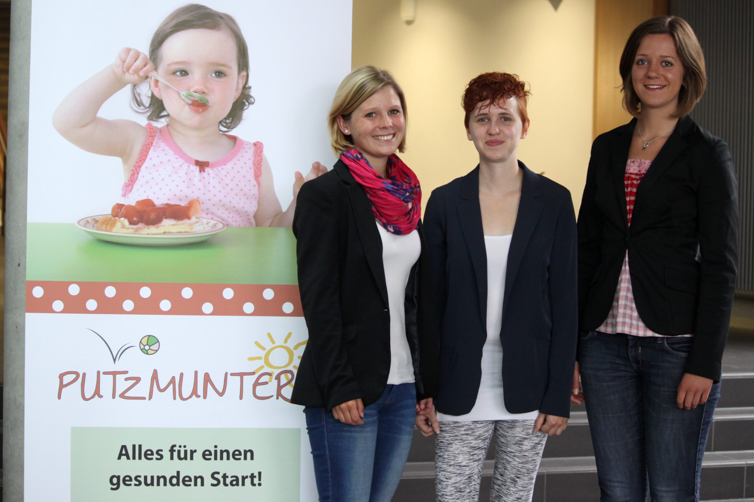 Drei Frauen der Hochschule Coburg stehen neben einem Banner, auf dem ein Spaghetti essendes Kind abgebildet ist. Auf dem Banner steht „PUTZMUNTER“ und „Alles für einen gesunden Start!“ Sie lächeln herzlich, sind leger gekleidet und verkörpern den Geist eines gesunden Starts.