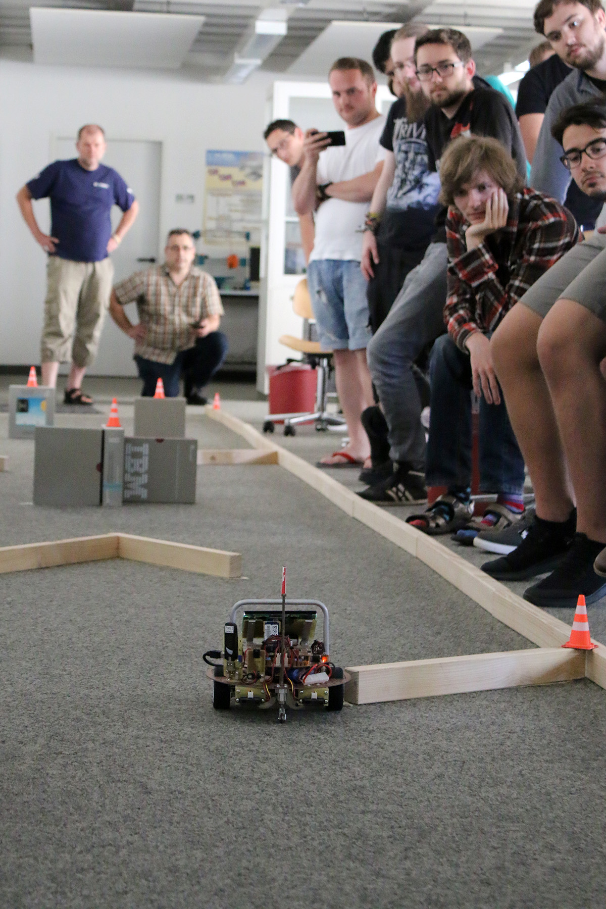 Eine Gruppe von Menschen an der Hochschule Coburg beobachtet, wie ein kleiner Roboter einen von Holzbarrieren und orangefarbenen Pylonen umgebenen Parcours befährt. Einige sitzen, andere stehen und alle beobachten die Bewegungen des Roboters mit großem Interesse. Die Kulisse scheint eine Indoor-Veranstaltung oder ein Workshop zu sein.