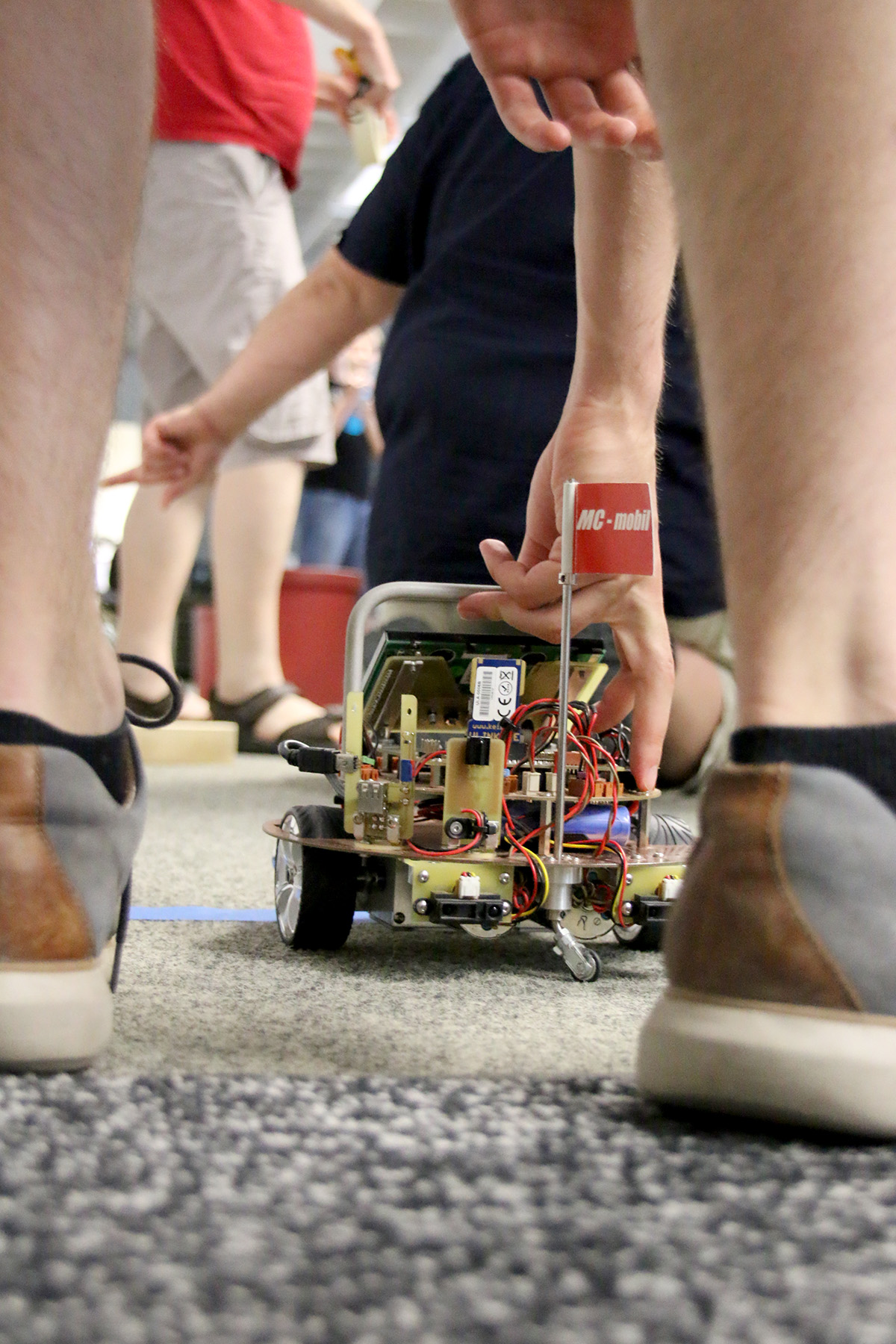 Eine Person kniet, um einen kleinen Roboter auf dem Boden einzustellen, umgeben von anderen in Freizeitkleidung an der Hochschule Coburg. Der Roboter mit sichtbaren Kabeln und Komponenten steht neben einer blauen Linie und auf einer kleinen Flagge steht „AC-motiv“.