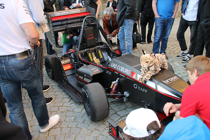 Ein Rennwagen mit der Aufschrift "UAS Coburg" steht auf einer Kopfsteinpflasterstraße umringt von einer Menschengruppe. Auf der Motorhaube sitzt ein Stofftier, während Beobachter, darunter Studierende der Hochschule Coburg, Fotos von der Szene machen.