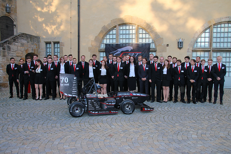 Eine Gruppe von Menschen in formeller Kleidung steht im Freien vor einem Gebäude der Hochschule Coburg, vor ihnen ein kleiner Rennwagen. Sie posieren für ein Gruppenfoto, dahinter ein Logo mit der Zahl 70.