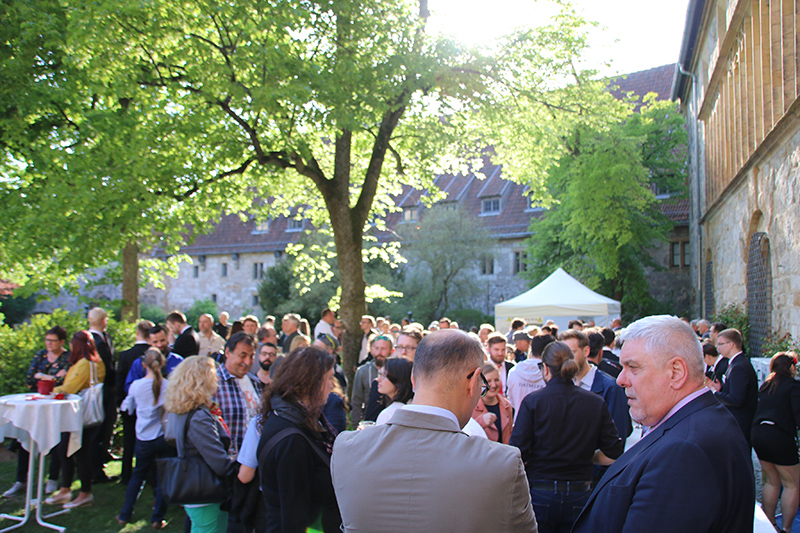 Eine große Gruppe von Menschen versammelte sich im Freien unter einem Baum und unterhielt sich während einer sonnigen Veranstaltung an der Hochschule Coburg. Ein weißes Zelt und ein historisches Steingebäude im Hintergrund bieten eine malerische Kulisse für zwangloses Beisammensein, umgeben von einigen Tischen.