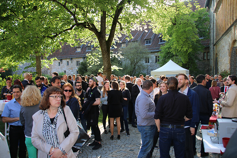 Eine große Gruppe von Menschen trifft sich unter einem Baum im Freien an der Hochschule Coburg. Sie sitzen an Tischen mit Getränken und Snacks. Im Hintergrund ist ein weißes Zelt zu sehen und unter dem klaren Himmel ist ein Gebäude mit roten Ziegeldächern zu erkennen.