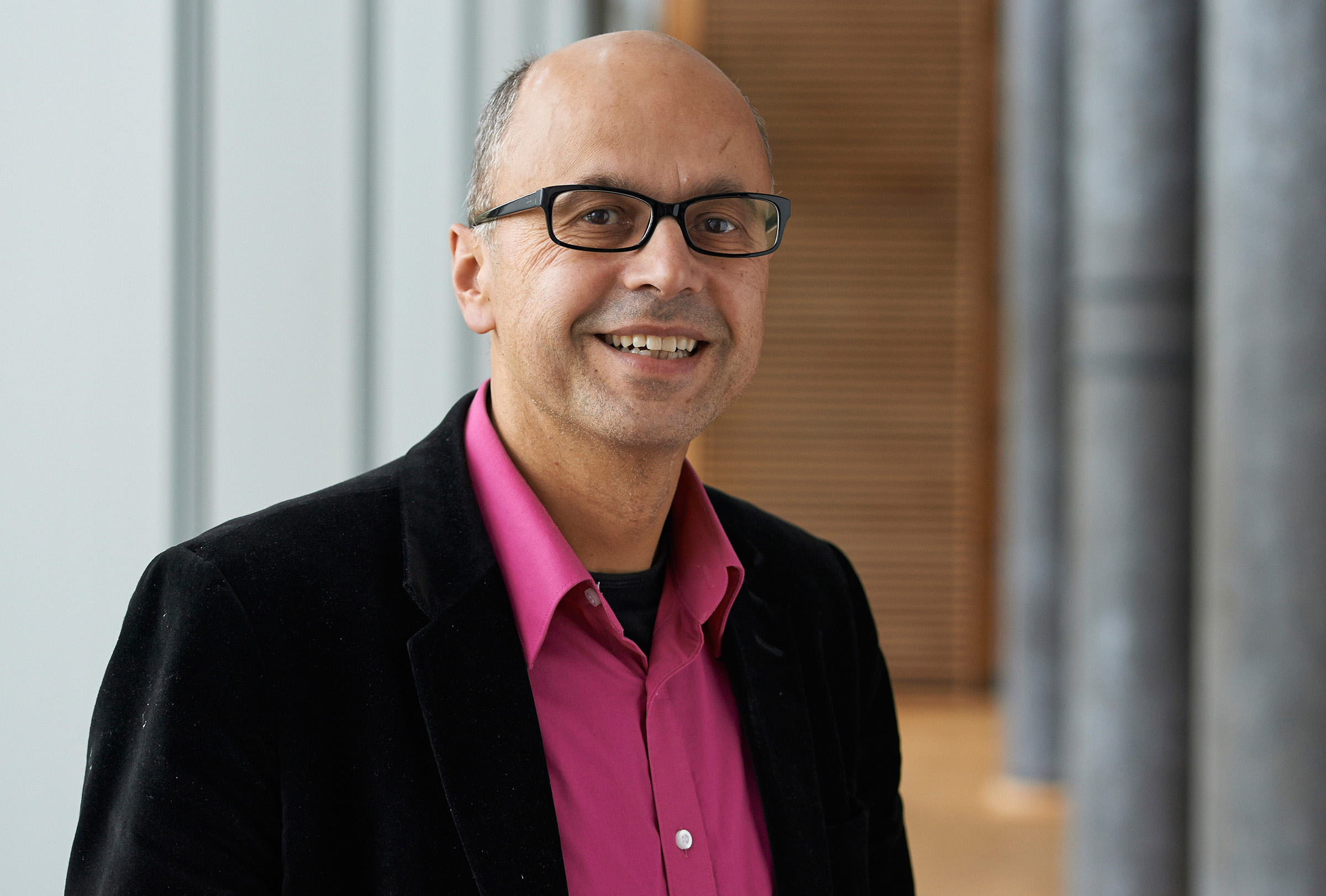 Eine lächelnde Person mit Brille, leuchtend rosa Hemd und schwarzem Blazer steht vor der modernen Architekturkulisse der Hochschule Coburg mit vertikalen Linien und Holzakzenten.