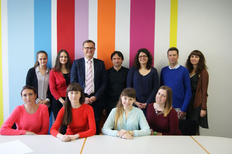 Eine Gruppe von zwölf Personen, darunter Männer und Frauen, posiert gemeinsam in einem Konferenzraum der Hochschule Coburg. Sie stehen und sitzen vor einer Wand mit vertikalen Streifen in leuchtenden Farben: Blau, Rosa, Orange und Rot. Einige sitzen an einem Tisch und tragen sowohl Freizeit- als auch Abendkleidung.