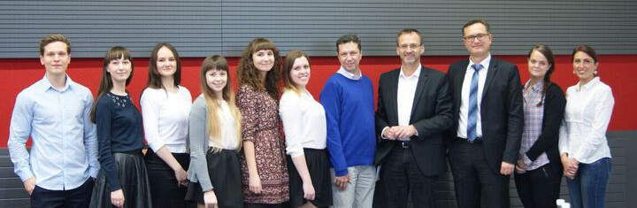 Eine Gruppe von elf Personen steht in einer Reihe und posiert für ein Foto an der Hochschule Coburg. Sie tragen formelle und halbformelle Kleidung, einige lächeln. Der Hintergrund besteht aus horizontalen grauen und roten Paneelen.