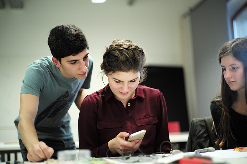 Drei junge Leute sitzen um einen Tisch in einem Studienraum der Hochschule Coburg. Einer steht und beugt sich vor, zwei sitzen. Die Person in der Mitte schaut auf ein Smartphone, die anderen schauen aufmerksam zu.