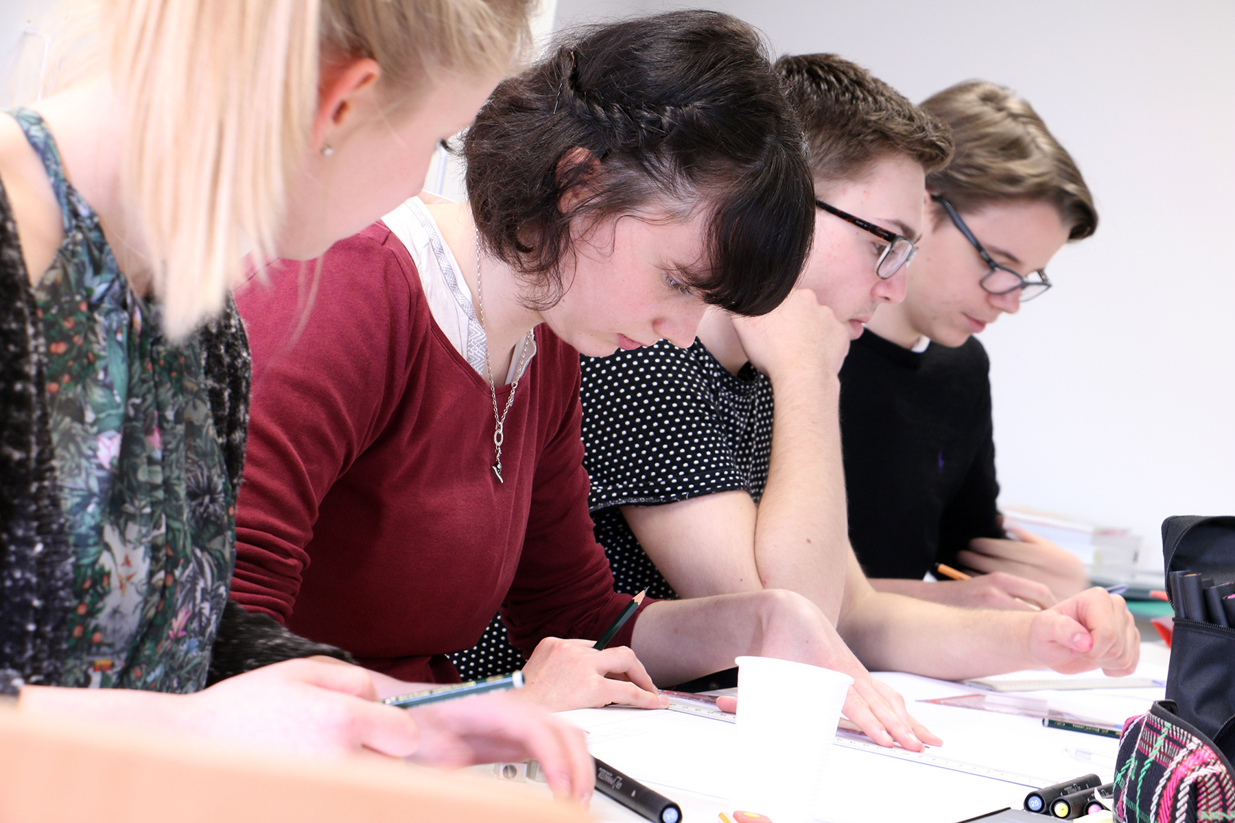 Vier Studierende sitzen in einer Reihe an einem Tisch und sind in ihre Studien vertieft. Sie machen sich Notizen und lesen aus den vor ihnen ausgebreiteten Materialien vor. Sie verkörpern damit die Hingabe, die man an der Hochschule Coburg oft sieht. Die Umgebung erinnert an ein Klassenzimmer oder eine Lerngruppe.