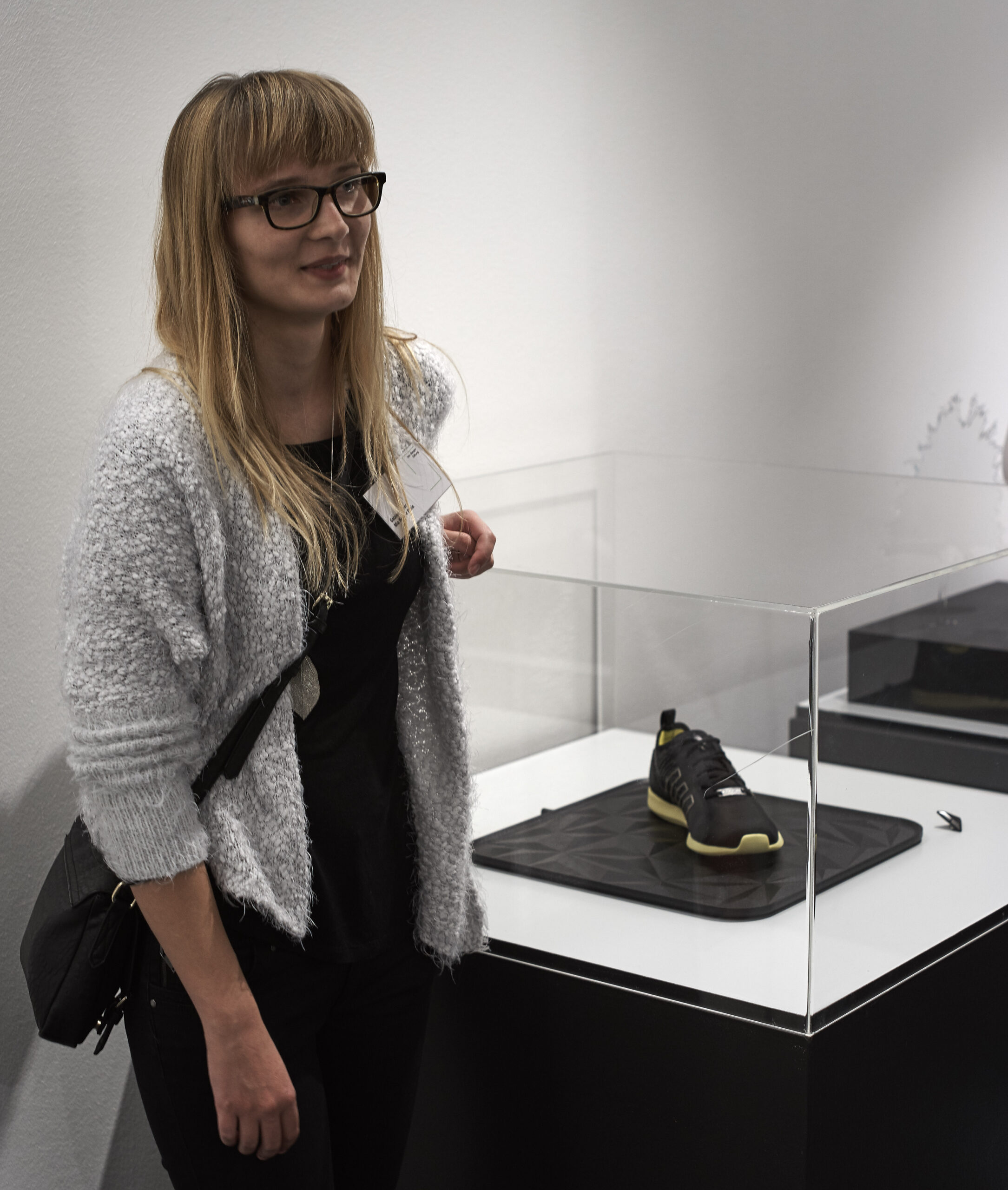 Eine Frau mit Brille und leichtem Pullover steht neben einer Vitrine mit einem schwarz-gelben Sneaker. Das minimalistische Setup, das an das moderne Designethos der Hochschule Coburg erinnert, ergänzt die klare Ästhetik der weißen Wand.