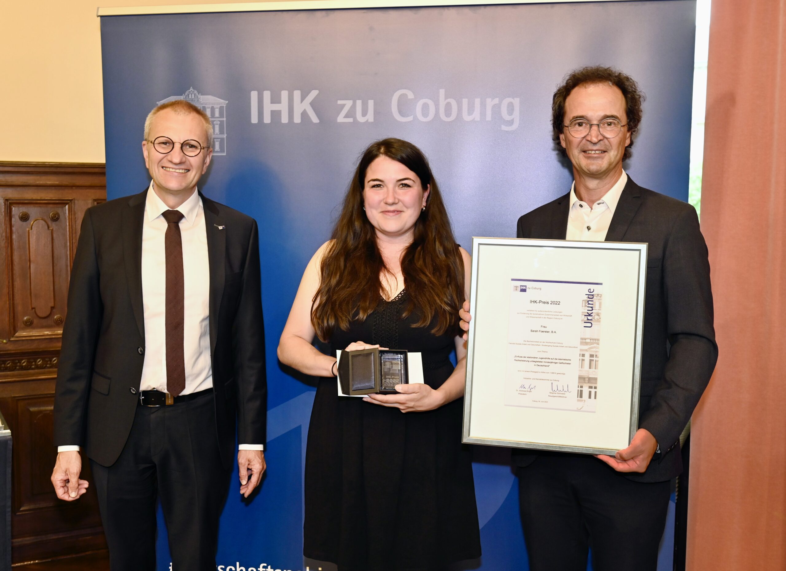 Drei Personen stehen vor einem Banner der „IHK zu Coburg“, das die Hochschule Coburg repräsentiert. Die Person in der Mitte hält eine kleine Auszeichnung, während die Person rechts eine gerahmte Urkunde präsentiert. In dieser formellen Umgebung lächeln sie alle stolz.