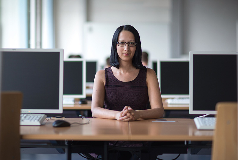 Eine Frau mit Brille und langen dunklen Haaren sitzt an einem Schreibtisch und blickt in die Kamera. Sie ist umgeben von Computermonitoren in einem hellen, modernen Büro im hochmodernen Computerlabor der Hochschule Coburg.