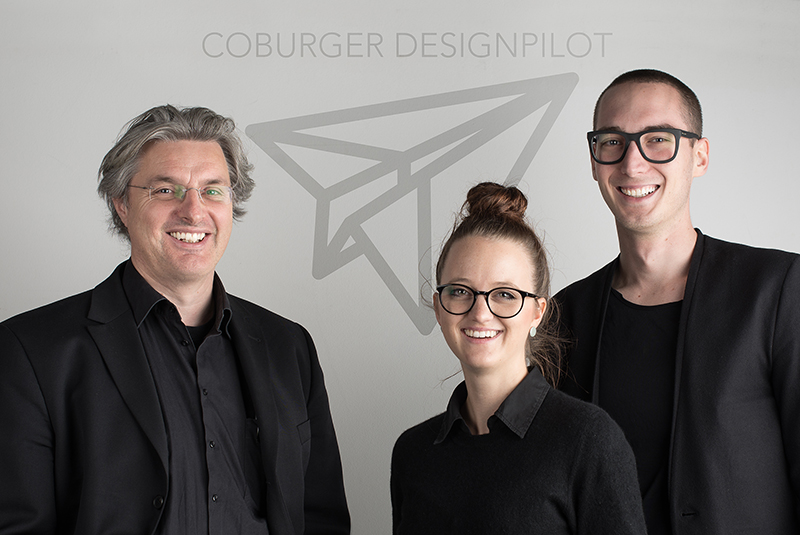 Drei lächelnde Menschen in schwarzer Kleidung stehen vor einer Wand mit dem Logo „COBURGER DESIGNPILOT“. Das Design eines Papierfliegers steht für den Innovationsgeist der Hochschule Coburg.