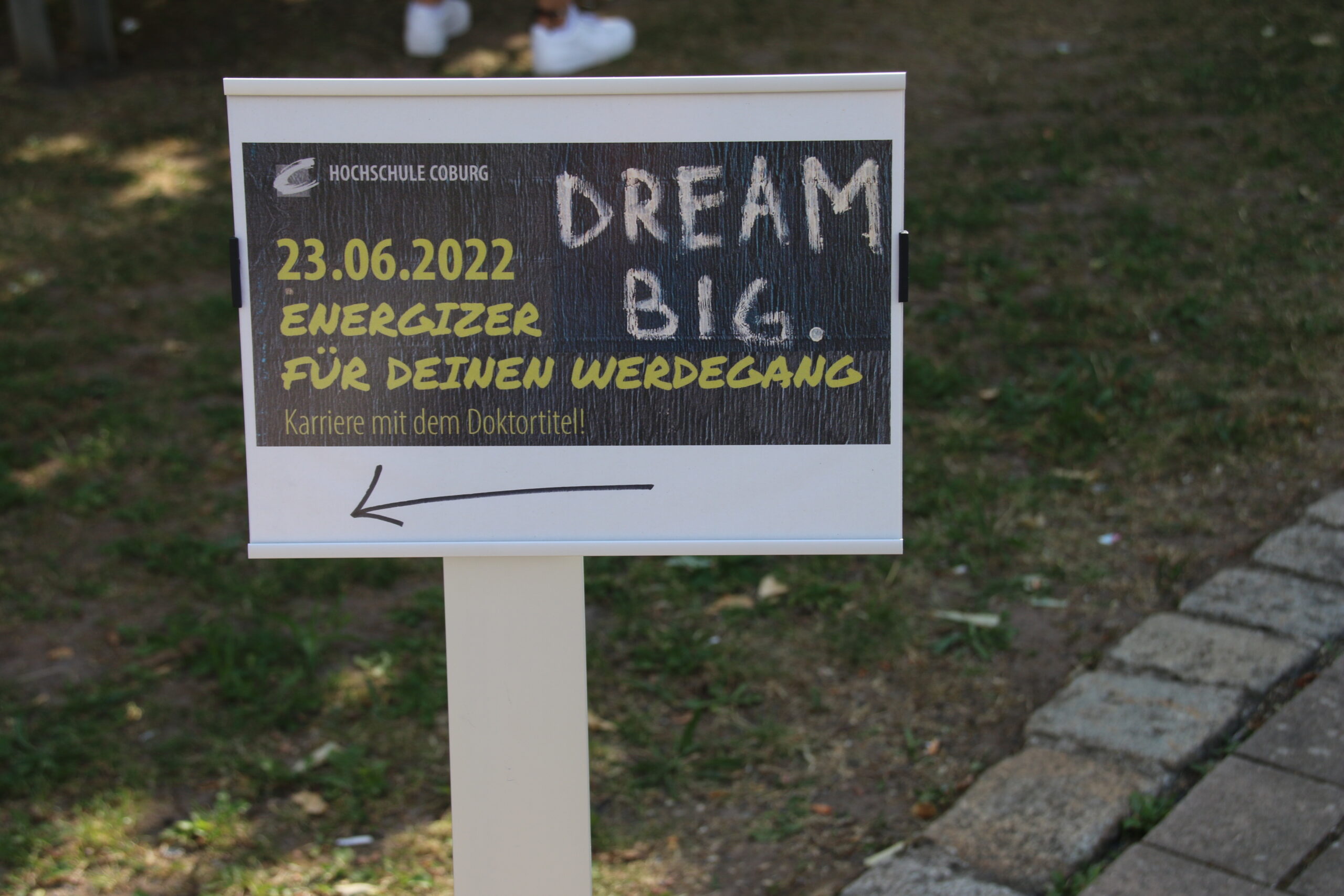 Auf einem Schild auf dem Rasen steht auf Deutsch: „Hochschule Coburg. 23.06.2022. Energizer für deinen Werdegang. Karriere mit dem Doktortitel!“ mit „Dream Big“ kühn mit Kreide skizziert und einem Pfeil, der nach links auf die Möglichkeiten der Hochschule Coburg verweist.