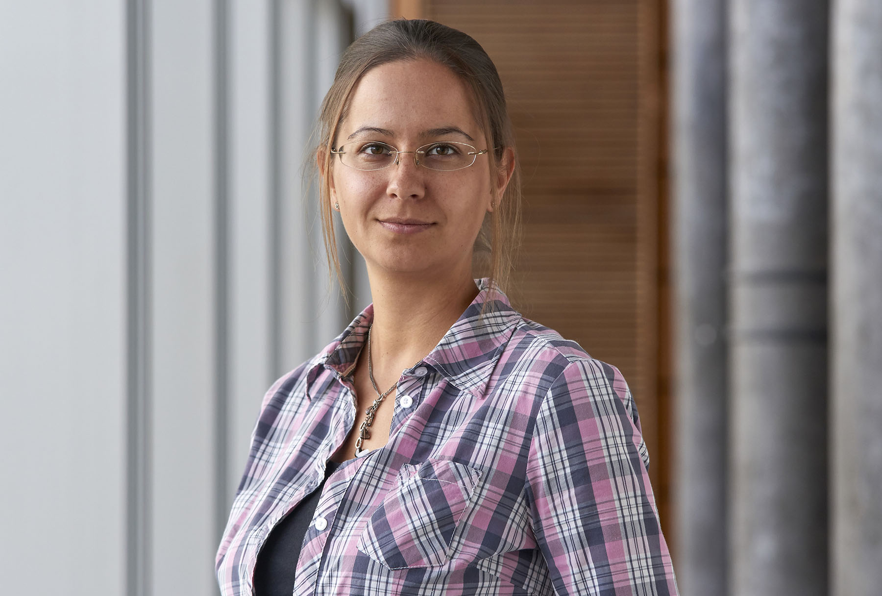 Eine Frau mit zurückgebundenem langem Haar trägt eine Brille und ein kariertes Hemd. Sie steht in einem Raum neben einem großen Fenster und blickt mit neutralem Gesichtsausdruck direkt in die Kamera. Vielleicht denkt sie über ihre Zeit an der Hochschule Coburg nach.