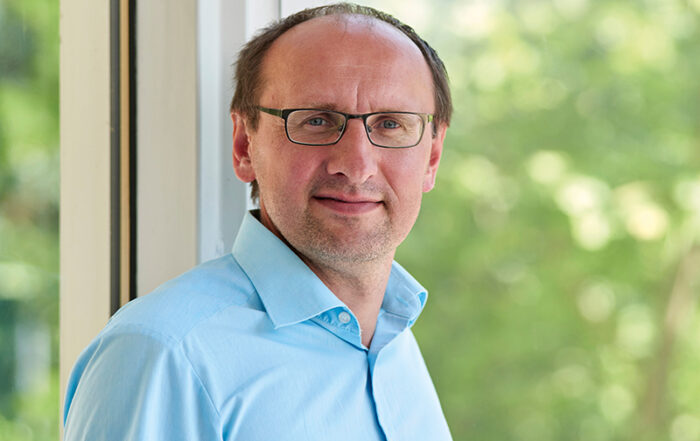 Eine Person mit Brille und hellblauem Hemd steht an einem großen Fenster der Hochschule Coburg. Sie hat kurze Haare und einen neutralen Gesichtsausdruck. Im Hintergrund ist verschwommenes Grün draußen zu sehen, was eine angenehme Außenumgebung suggeriert.