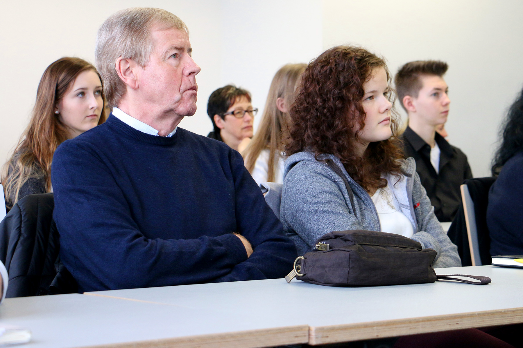 In einem Hörsaal der Hochschule Coburg hört eine Gruppe von Menschen unterschiedlichen Alters aufmerksam zu. Der ältere Mann in der ersten Reihe trägt einen dunklen Pullover, neben ihm eine junge Frau mit Locken und grauer Jacke. Im Hintergrund sind weitere zu sehen, die konzentriert sitzen.