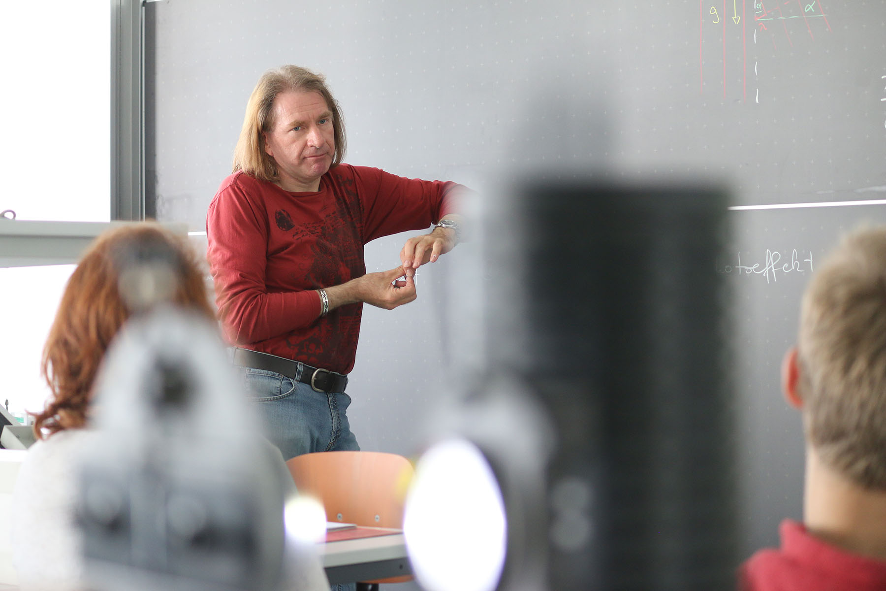 Eine Person mit langen Haaren und rotem Hemd steht vor einer Tafel und gestikuliert, während sie an der Hochschule Coburg unterrichtet. Im Vordergrund sind verschwommene Studenten und eine Lichtquelle zu sehen, inmitten von Diagrammen und Notizen an der Tafel, die die akademische Kulisse vervollständigen.