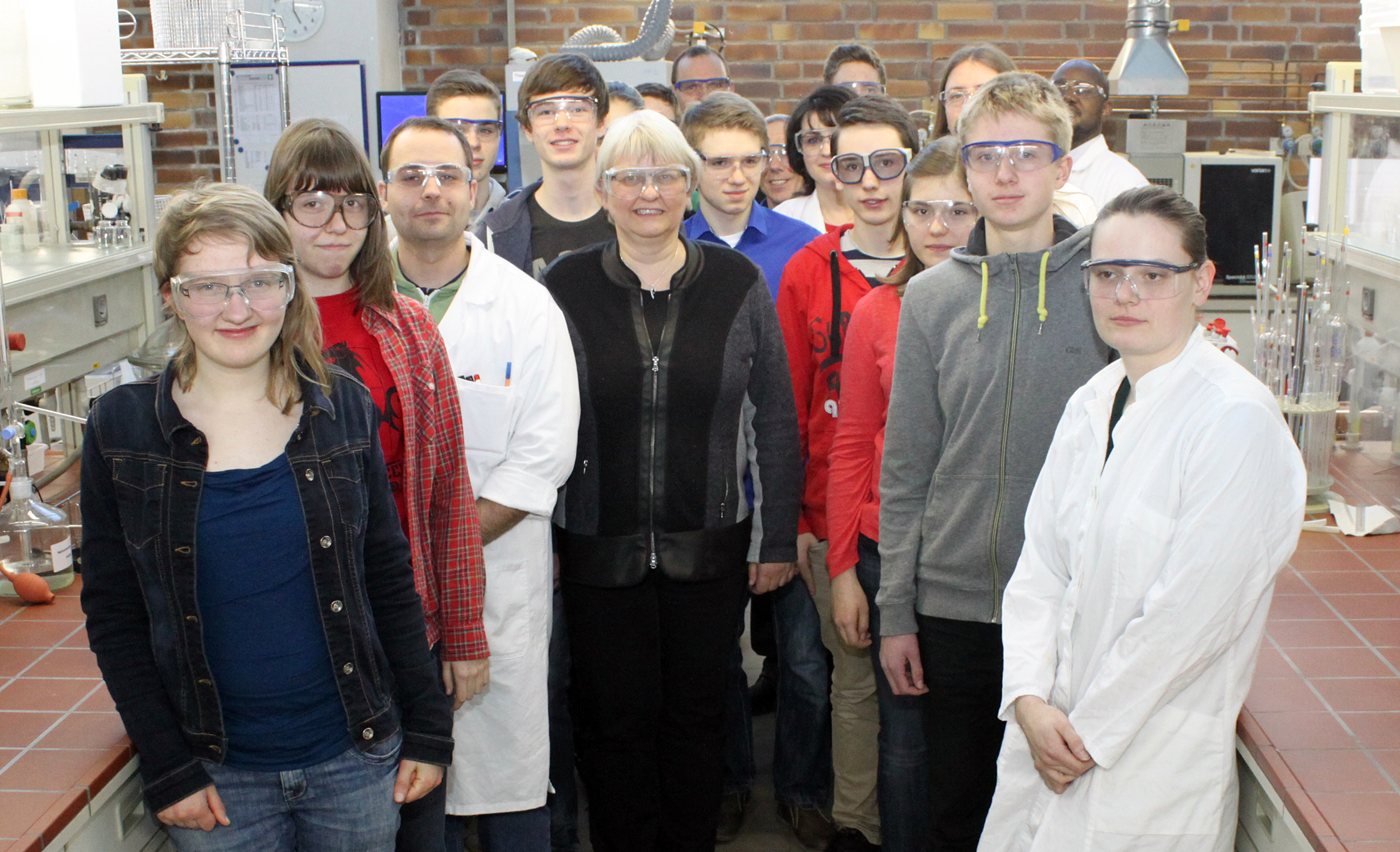 Eine Gruppe von Menschen mit Schutzbrillen und Laborkitteln steht in einem Chemielabor der Hochschule Coburg. Sie posieren für ein Foto vor Labortischen und -geräten, im Hintergrund sind Backsteinwände zu sehen.