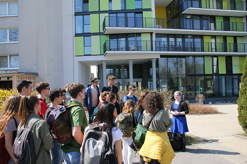 Eine Gruppe von Studierenden steht vor der Hochschule Coburg, einem modernen Gebäude mit grünen und gelben Tafeln. Aufmerksam hören sie einem Vortragenden zu und machen sich Notizen. Es ist ein sonniger Tag.