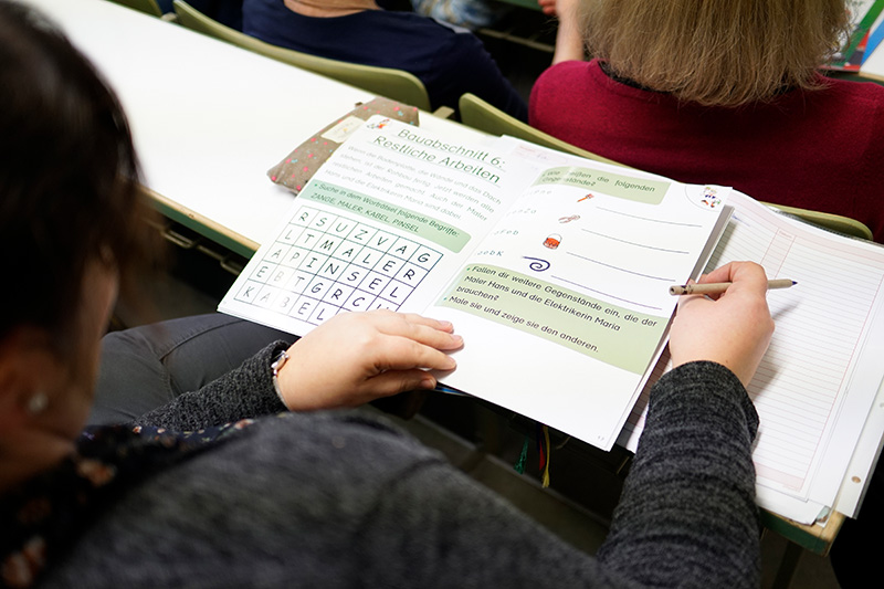 In einem Hörsaal der Hochschule Coburg sitzt eine Person mit einem aufgeschlagenen Buch in Deutsch auf dem Schoß, in dem ein Wortsuchrätsel steht. Die Mitschüler in der Nähe konzentrieren sich auf ihre Studien, während auf dem Tisch neben ihnen ein Notizblock liegt.