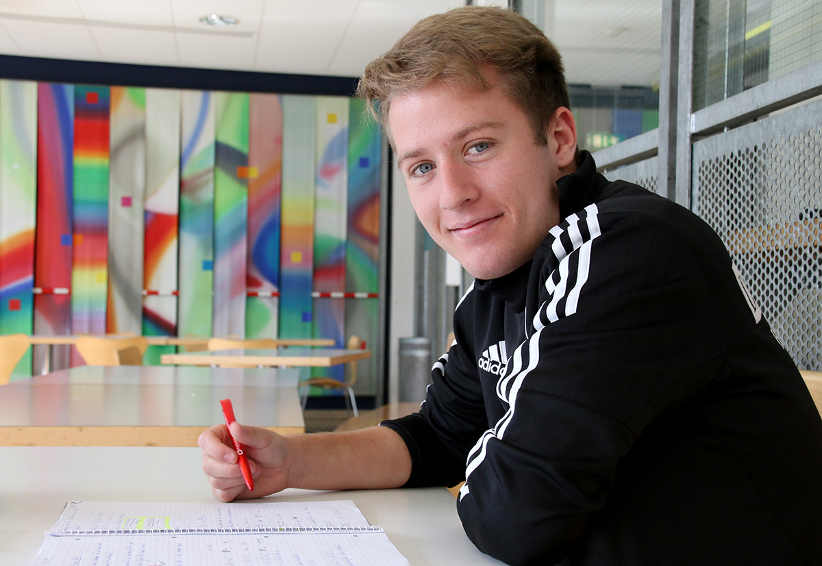 Ein junger Mensch mit kurzen Haaren sitzt an einem Tisch, hält einen roten Stift in der Hand und blickt in die Kamera. Er trägt eine schwarz-weiße Jacke, auf die dezent das Emblem der Hochschule Coburg gestickt ist. Im Hintergrund ist farbenfrohe abstrakte Kunst zu sehen, die der Szene Lebendigkeit verleiht.